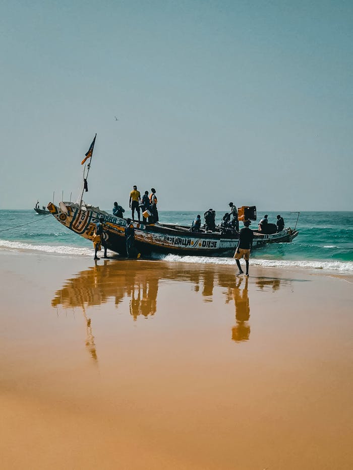 men-standing-on-boat-docked-in-shallow-sea-water-11385115 Colorful wooden fishing boat with crew on Kayar Beach, Senegal reflecting on sand under clear sky.